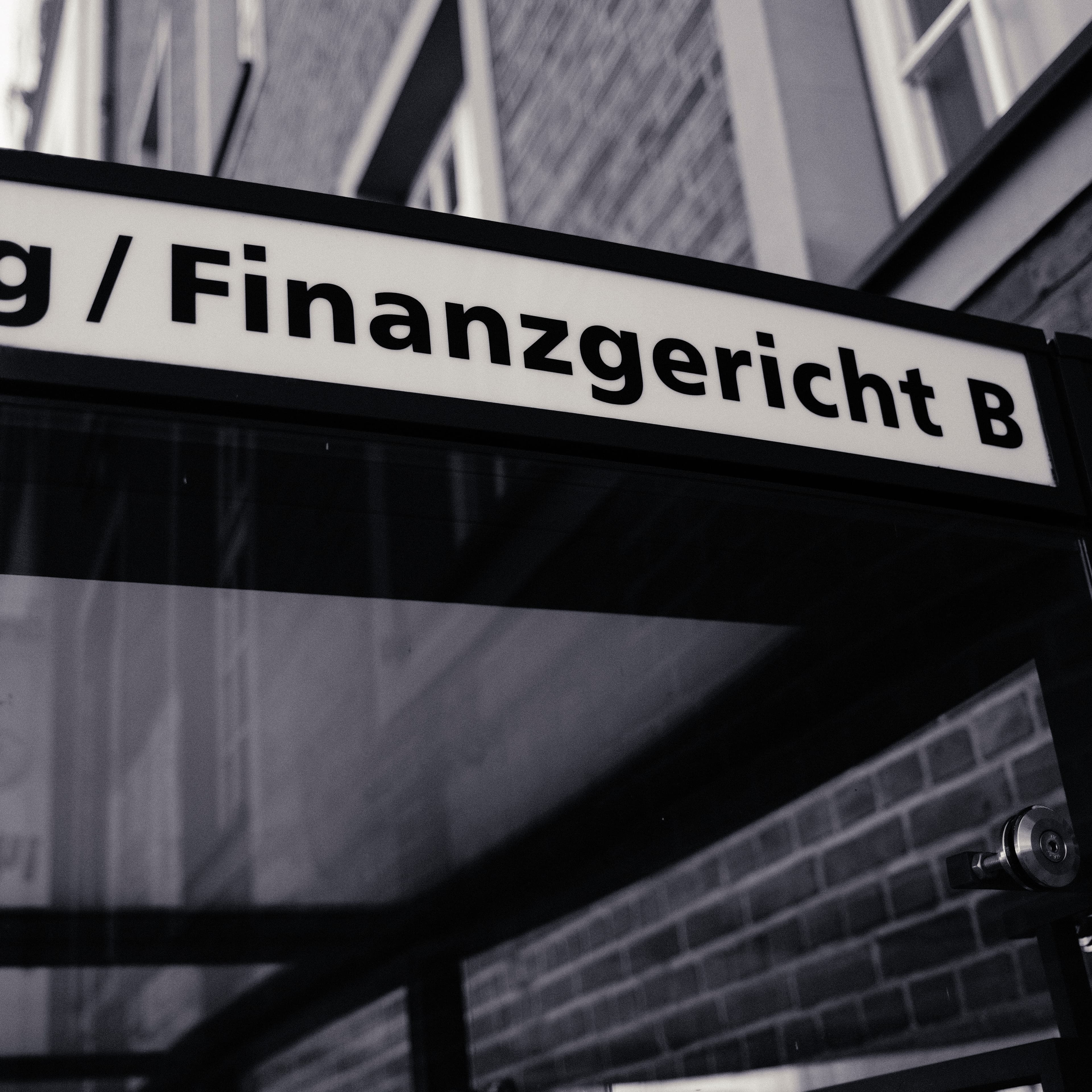 Beleuchtetes Hinweisschild mit der Aufschrift „Finanzgericht B“, von unten fotografiert.