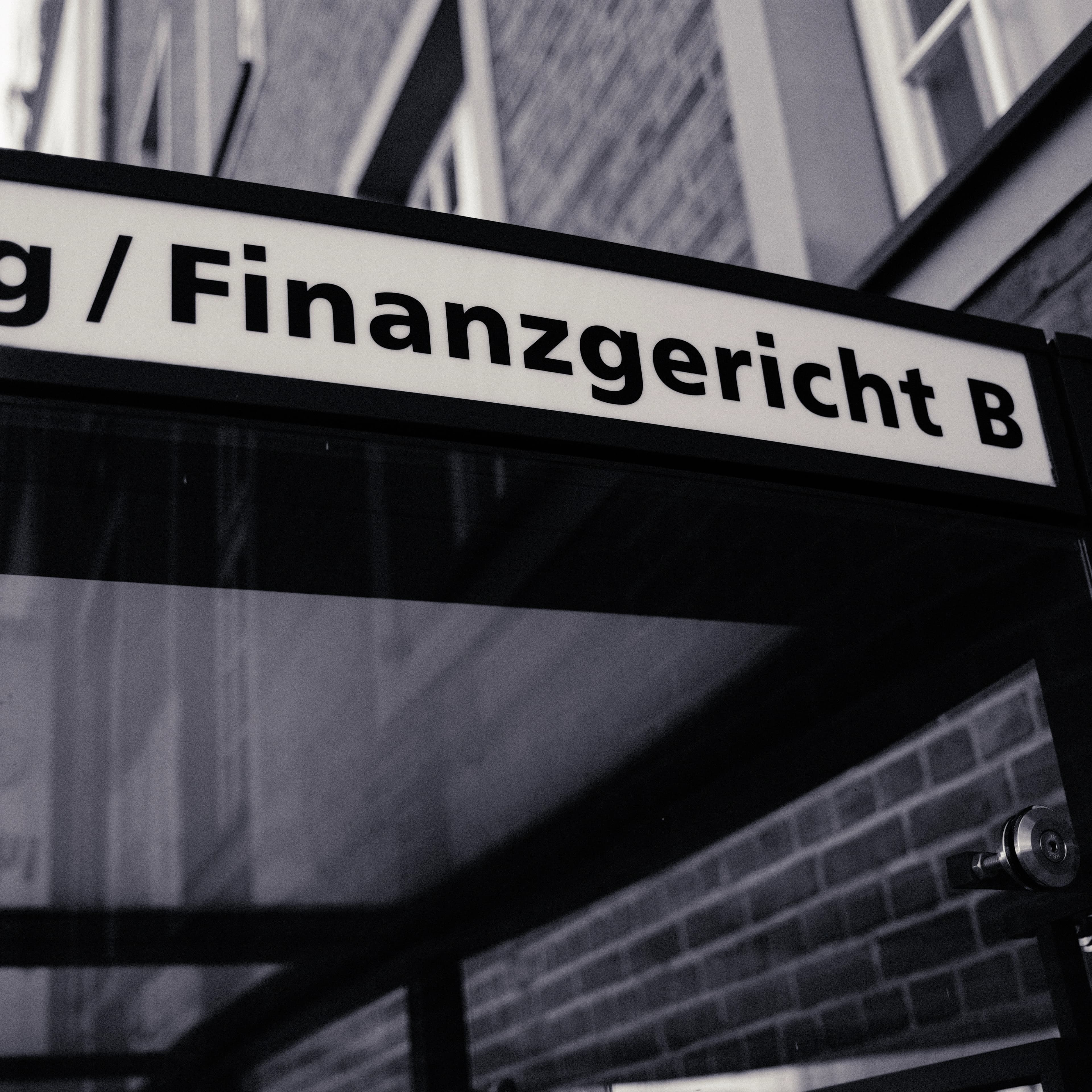 Beleuchtetes Hinweisschild mit der Aufschrift „Finanzgericht B“, von unten fotografiert.